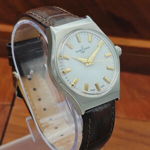 May include: A silver Ulysse Nardin wristwatch with a white face and gold hands and markers. It features a brown leather strap. The watch face displays the brand name and the word "SWISS".