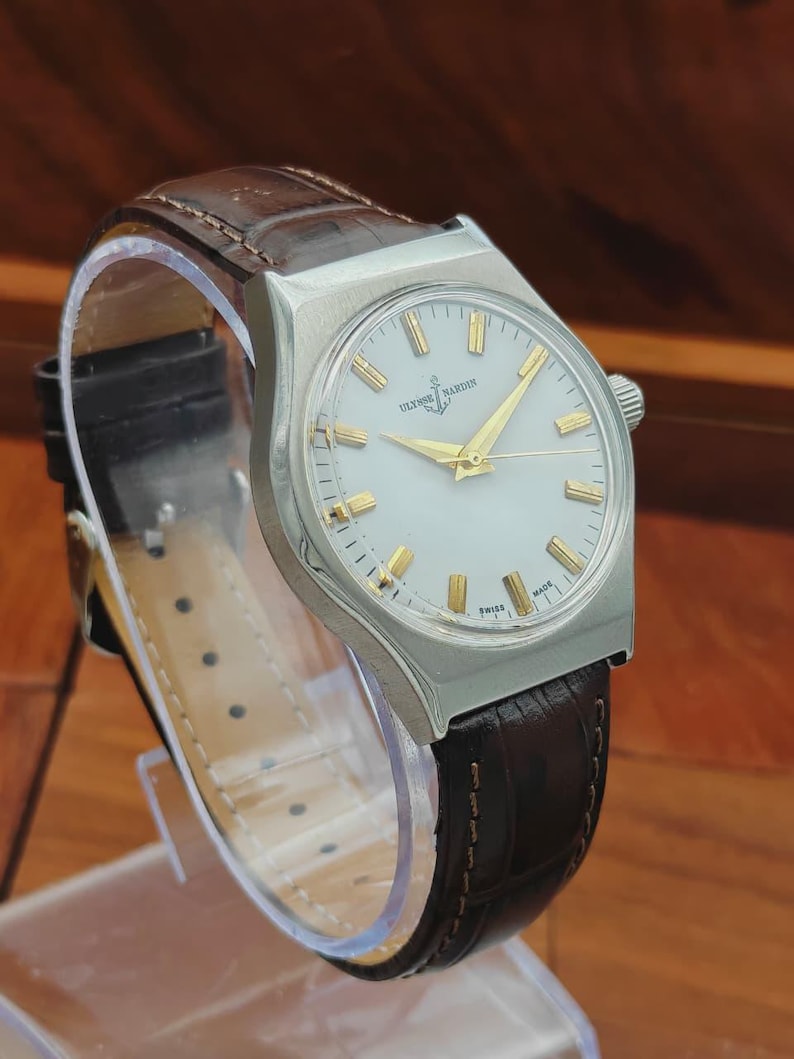 May include: A silver Ulysse Nardin wristwatch with a white face and gold hands and markers. It features a brown leather strap. The watch face displays the brand name and the word "SWISS".