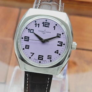 Puede incluir: Un reloj de pulsera Ulysse Nardin plateado con esfera lavanda y agujas negras. El reloj tiene n&uacute;meros negros y una correa de cuero marr&oacute;n. La esfera del reloj muestra el logotipo de Ulysse Nardin y las palabras "SWISS MADE".