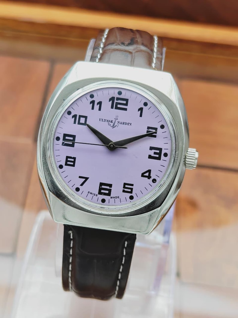 Puede incluir: Un reloj de pulsera Ulysse Nardin plateado con esfera lavanda y agujas negras. El reloj tiene n&uacute;meros negros y una correa de cuero marr&oacute;n. La esfera del reloj muestra el logotipo de Ulysse Nardin y las palabras "SWISS MADE".