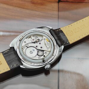 May include: A close-up view of the back of a silver-toned wristwatch, revealing its intricate internal mechanisms. The watch has a black leather strap. The watch's gears, springs, and other components are visible through the clear case back.