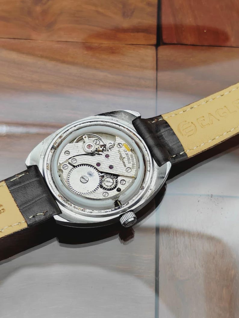 May include: A close-up view of the back of a silver-toned wristwatch, revealing its intricate internal mechanisms. The watch has a black leather strap. The watch's gears, springs, and other components are visible through the clear case back.