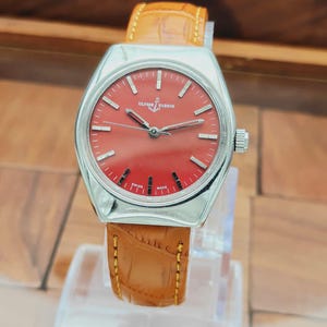 Puede incluir: Un reloj Ulysse Nardin vintage con esfera roja y caja plateada. El reloj presenta una correa de cuero marr&oacute;n con costuras amarillas. El reloj se exhibe sobre un soporte transparente.