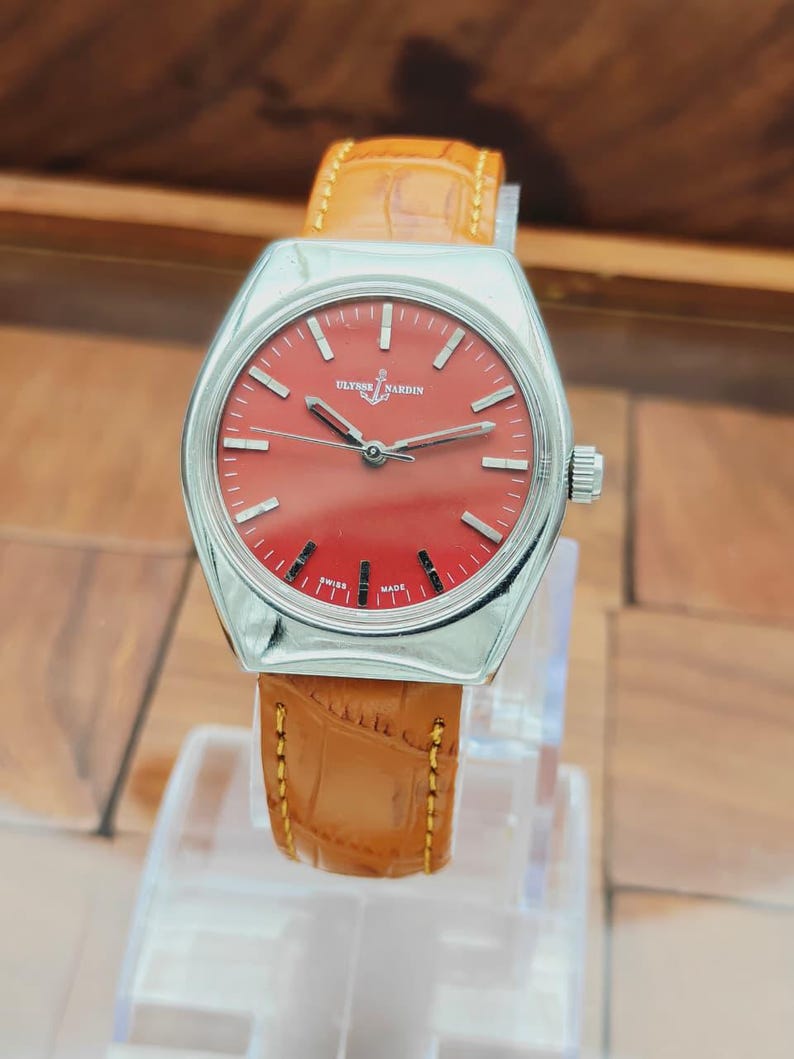 Puede incluir: Un reloj Ulysse Nardin vintage con esfera roja y caja plateada. El reloj presenta una correa de cuero marr&oacute;n con costuras amarillas. El reloj se exhibe sobre un soporte transparente.