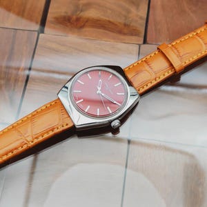 Puede incluir: Un reloj de pulsera vintage con esfera roja y caja plateada. El reloj tiene una correa de cuero marr&oacute;n claro con un patr&oacute;n de cocodrilo. El reloj est&aacute; sobre una superficie de madera.