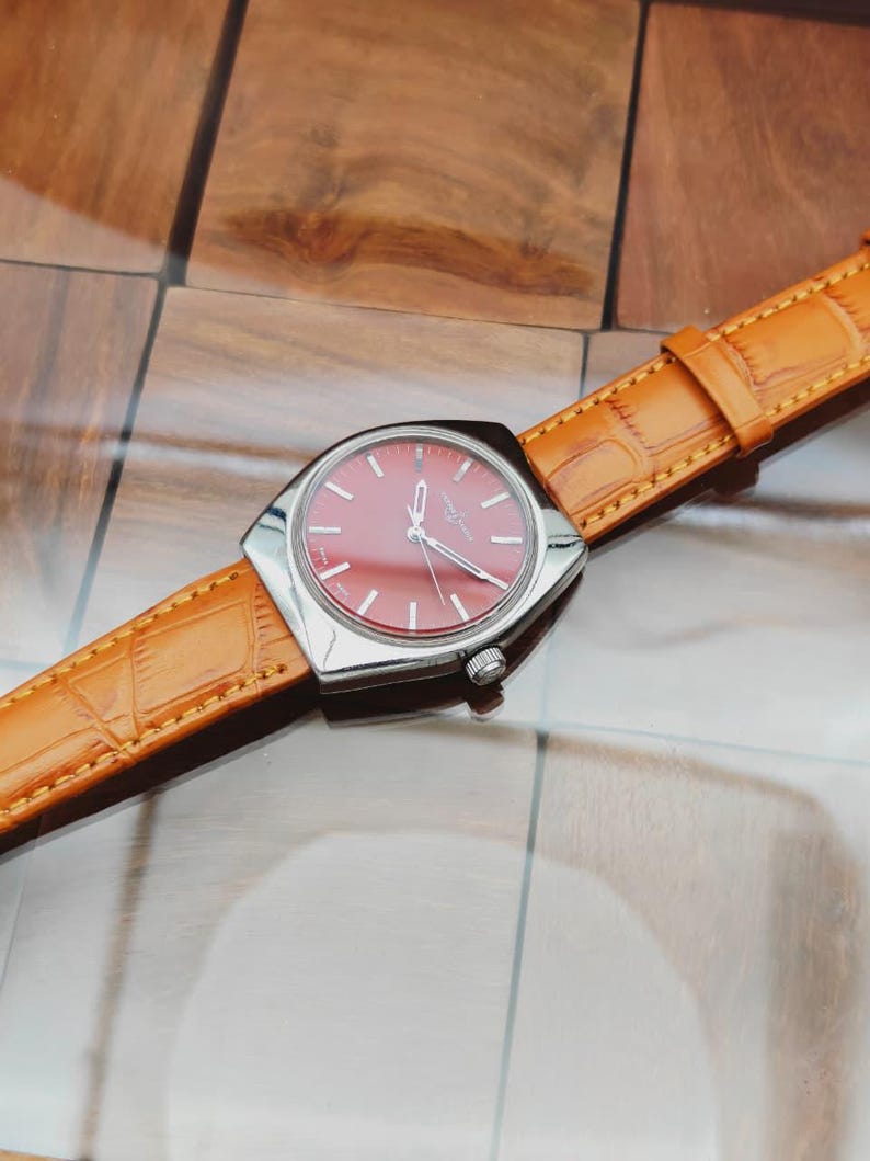 Puede incluir: Un reloj de pulsera vintage con esfera roja y caja plateada. El reloj tiene una correa de cuero marr&oacute;n claro con un patr&oacute;n de cocodrilo. El reloj est&aacute; sobre una superficie de madera.