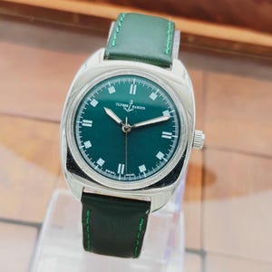 K&ouml;nnte beinhalten: Eine silberfarbene Armbanduhr mit gr&uuml;nem Zifferblatt und gr&uuml;nem Lederarmband. Das Zifferblatt hat wei&szlig;e Markierungen und Zeiger, mit dem Markennamen "Ulysse Nardin". Das Armband ist mit gr&uuml;nem Faden gen&auml;ht.