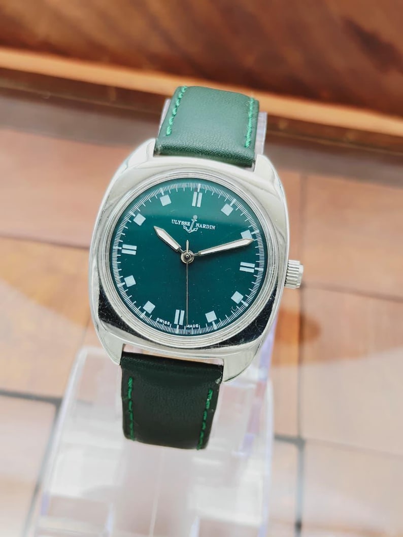 K&ouml;nnte beinhalten: Eine silberfarbene Armbanduhr mit gr&uuml;nem Zifferblatt und gr&uuml;nem Lederarmband. Das Zifferblatt hat wei&szlig;e Markierungen und Zeiger, mit dem Markennamen "Ulysse Nardin". Das Armband ist mit gr&uuml;nem Faden gen&auml;ht.