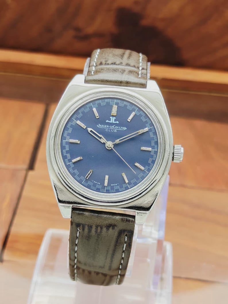 Puede incluir: Un reloj Jaeger-LeCoultre Club plateado con esfera azul oscuro y correa de cuero marr&oacute;n. El reloj tiene marcadores de hora y agujas plateadas, y la correa est&aacute; cosida con hilo blanco. La esfera del reloj dice "Jaeger-LeCoultre Club".