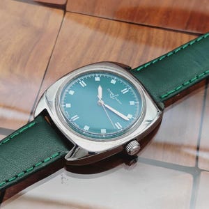 K&ouml;nnte beinhalten: Eine Vintage-Armbanduhr mit t&uuml;rkisblauem Zifferblatt und silbernem Geh&auml;use. Die Uhr hat ein gr&uuml;nes Lederarmband mit wei&szlig;en N&auml;hten. Das Zifferblatt zeigt den Markennamen "Ulysse Nardin".
