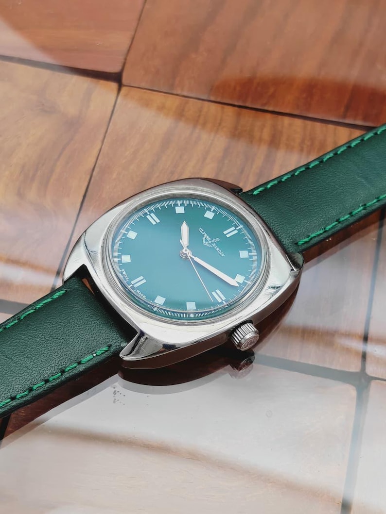 K&ouml;nnte beinhalten: Eine Vintage-Armbanduhr mit t&uuml;rkisblauem Zifferblatt und silbernem Geh&auml;use. Die Uhr hat ein gr&uuml;nes Lederarmband mit wei&szlig;en N&auml;hten. Das Zifferblatt zeigt den Markennamen "Ulysse Nardin".