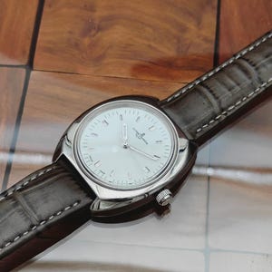 May include: A silver-toned wristwatch with a white face and a brown leather band. The watch has a round face with silver hands and markers. The band is stitched with white thread.