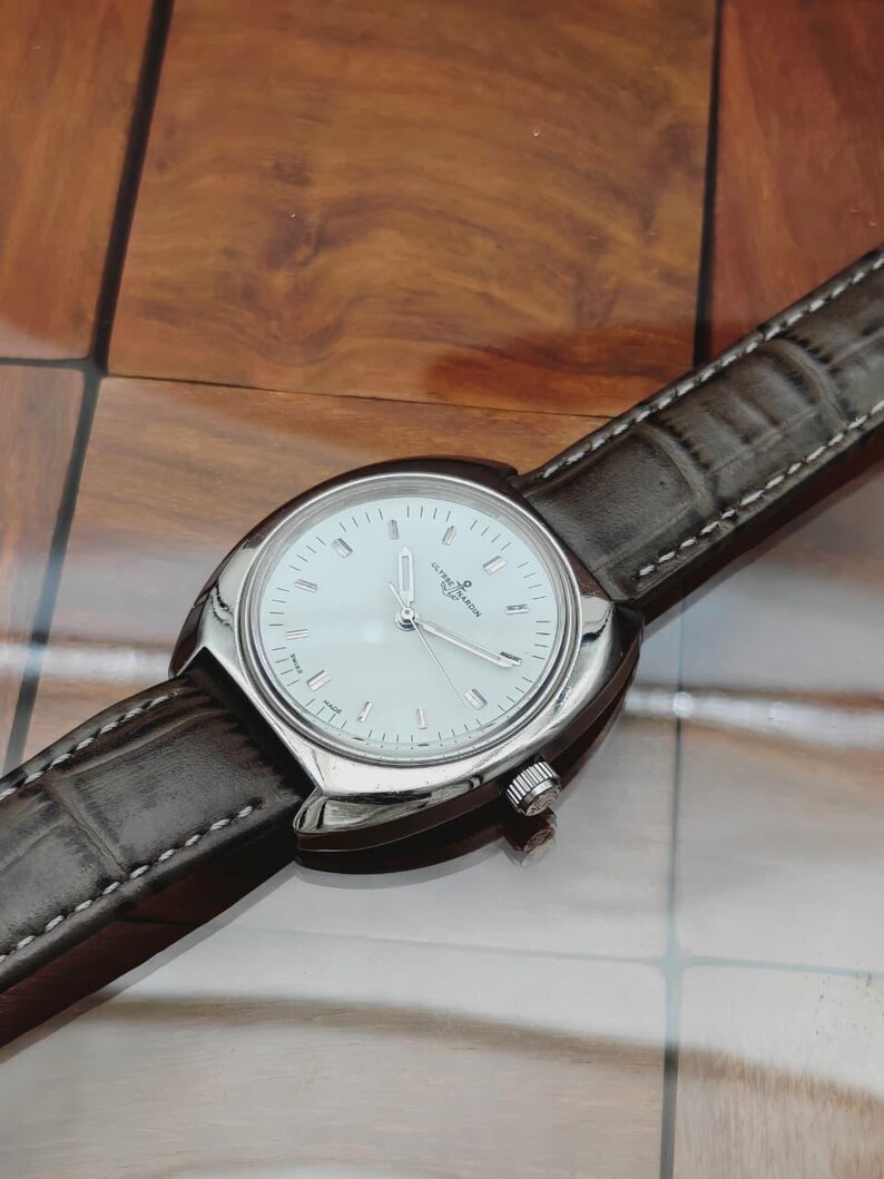 May include: A silver-toned wristwatch with a white face and a brown leather band. The watch has a round face with silver hands and markers. The band is stitched with white thread.