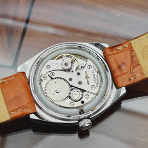 Puede incluir: Primer plano de un reloj de pulsera vintage con caja plateada y correa de cuero marr&oacute;n. El intrincado mecanismo interno del reloj es visible, mostrando engranajes, joyas y el nombre de la marca. Fondo neutro.
