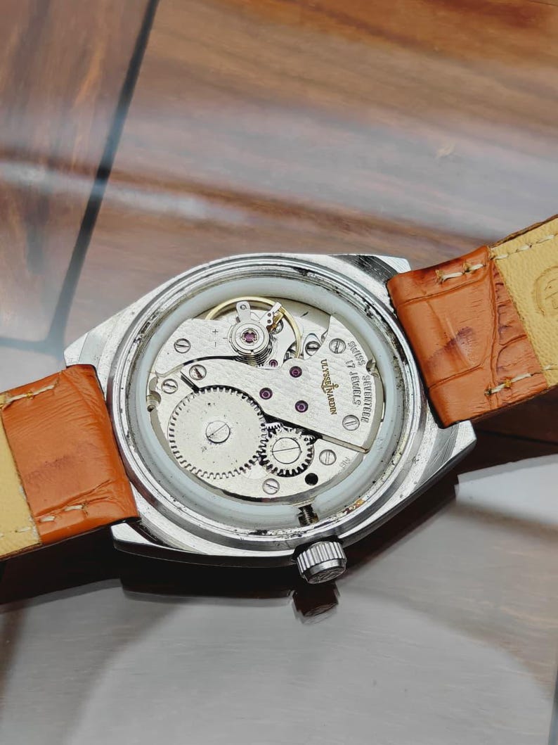 Puede incluir: Primer plano de un reloj de pulsera vintage con caja plateada y correa de cuero marr&oacute;n. El intrincado mecanismo interno del reloj es visible, mostrando engranajes, joyas y el nombre de la marca. Fondo neutro.