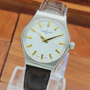 May include: A silver wristwatch with a white face, gold hands and markers, and a brown leather strap. The watch face displays the brand name "Ulysse Nardin" and the words "Swiss Made". The watch has a classic, elegant design.