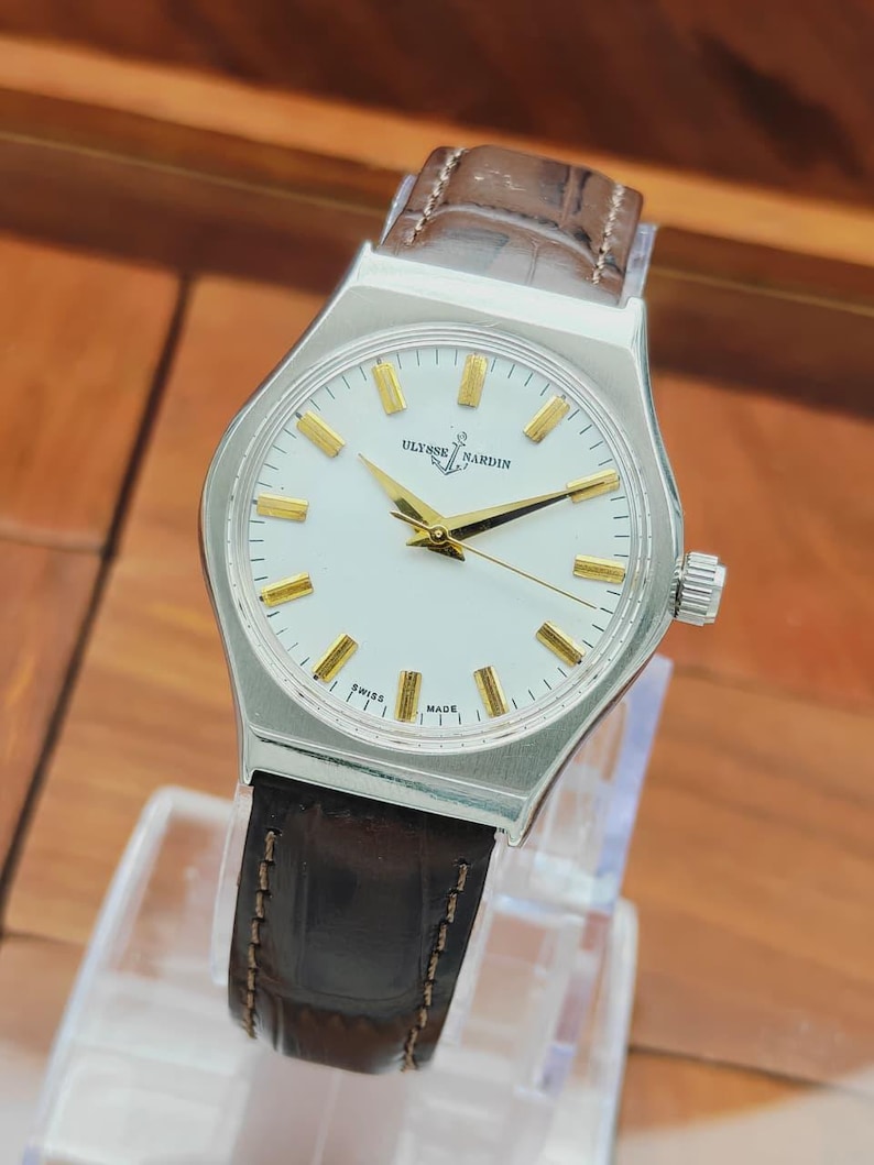 May include: A silver wristwatch with a white face, gold hands and markers, and a brown leather strap. The watch face displays the brand name "Ulysse Nardin" and the words "Swiss Made". The watch has a classic, elegant design.
