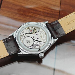 May include: A silver wristwatch with a transparent back, revealing the complex mechanical workings. The watch features a brown leather strap. The internal gears and components are visible. A classic timepiece.