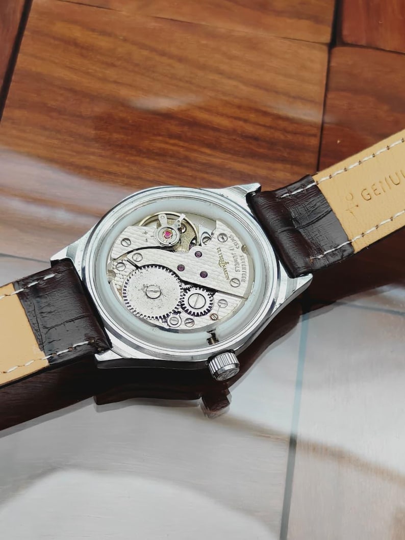 May include: A silver wristwatch with a transparent back, revealing the complex mechanical workings. The watch features a brown leather strap. The internal gears and components are visible. A classic timepiece.