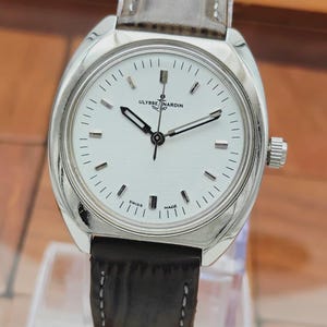 May include: A silver-toned Ulysse Nardin wristwatch with a white face and black hands. The watch has a brown leather band and a silver bezel. The watch face features the text "Ulysse Nardin" and "Swiss Made".