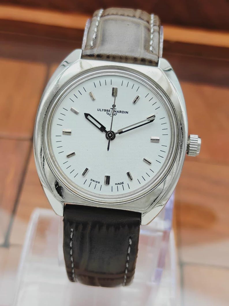 May include: A silver-toned Ulysse Nardin wristwatch with a white face and black hands. The watch has a brown leather band and a silver bezel. The watch face features the text "Ulysse Nardin" and "Swiss Made".