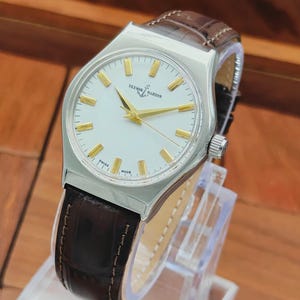 May include: A silver wristwatch with a white face, gold hands and markers, and a brown leather strap. The watch face displays the text "ULYSSE NARDIN" and "SWISS MADE". The watch is displayed on a clear stand.