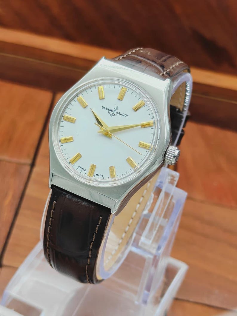 May include: A silver wristwatch with a white face, gold hands and markers, and a brown leather strap. The watch face displays the text "ULYSSE NARDIN" and "SWISS MADE". The watch is displayed on a clear stand.