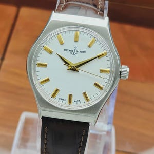 May include: A silver Ulysse Nardin wristwatch with a white face and gold hands and markers. The watch has a brown leather strap. The watch face displays the text "Ulysse Nardin" and "Swiss Made".