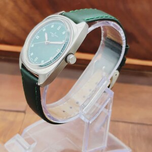 K&ouml;nnte beinhalten: Eine Vintage-Armbanduhr mit silbernem Geh&auml;use und gr&uuml;nem Zifferblatt. Die Uhr hat ein gr&uuml;nes Lederarmband und eine silberne Krone. Die Uhr wird auf einem transparenten Kunststoffst&auml;nder ausgestellt. Das Zifferblatt hat silberne Stundenmarkierungen und Zeiger.