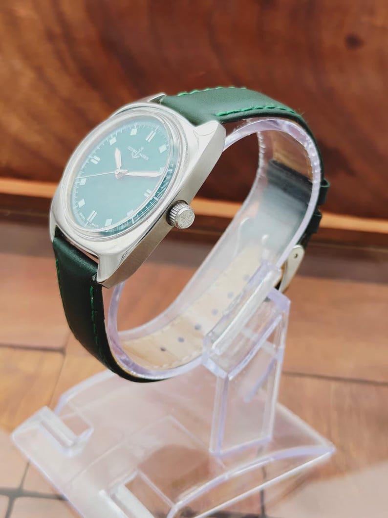 K&ouml;nnte beinhalten: Eine Vintage-Armbanduhr mit silbernem Geh&auml;use und gr&uuml;nem Zifferblatt. Die Uhr hat ein gr&uuml;nes Lederarmband und eine silberne Krone. Die Uhr wird auf einem transparenten Kunststoffst&auml;nder ausgestellt. Das Zifferblatt hat silberne Stundenmarkierungen und Zeiger.