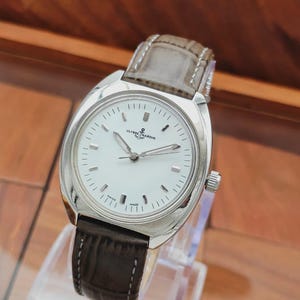 May include: A silver-toned wristwatch with a white face and a brown leather band. The watch face features black hour markers and the brand name "ULYSSE NARDIN". The watch is displayed on a clear stand.