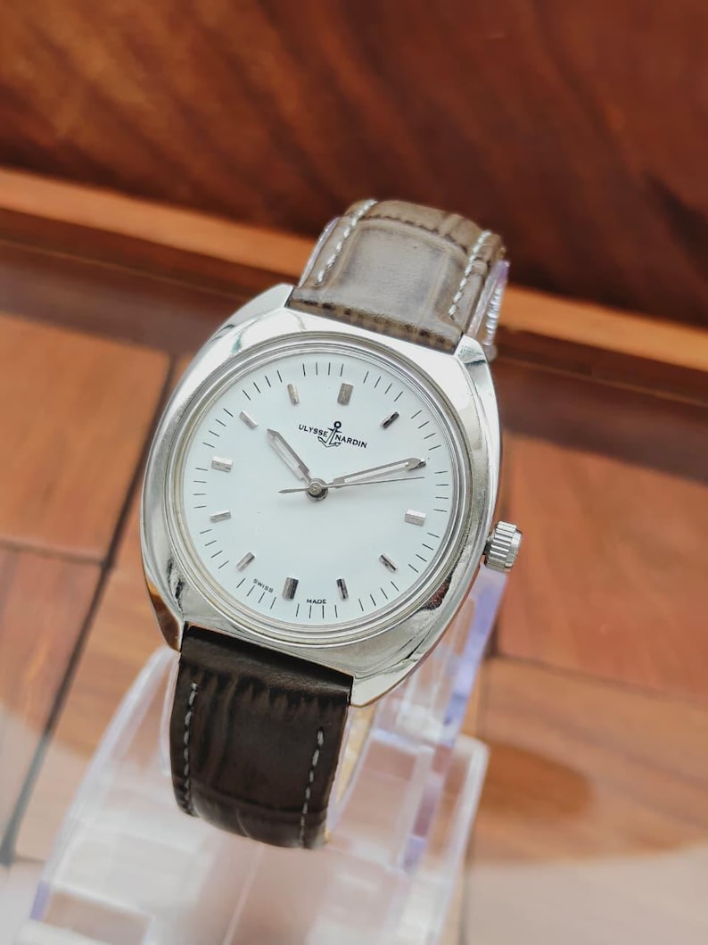 May include: A silver-toned wristwatch with a white face and a brown leather band. The watch face features black hour markers and the brand name "ULYSSE NARDIN". The watch is displayed on a clear stand.