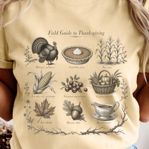 May include: A light yellow t-shirt featuring a dark brown graphic print titled Field Guide to Thanksgiving. The design displays illustrations of a turkey, pie, corn, berries, a basket, leaves, an acorn, and a gravy boat with botanical accents.