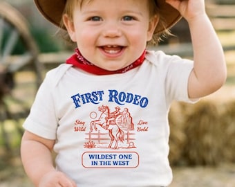 May include: A smiling baby wearing a brown cowboy hat, red bandana, blue jeans, and brown boots. The white onesie has the text "First Rodeo" and a graphic of a cowboy riding a horse. The baby is sitting on a wooden bench.