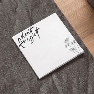Op de afbeelding: Een stapel witte plaknotities met de tekst "don't forget" in zwart schrift. De notities hebben ook een klein bloemmotief in de hoek. De notities liggen op een donkergrijze ondergrond, met een laptop en een pen in de buurt.