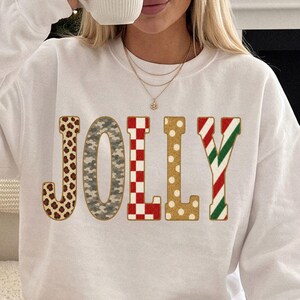May include: A white long-sleeve crewneck sweatshirt featuring the word JOLLY printed across the chest. Each letter has a distinct pattern: the J is leopard print, the O is grey camo, the L is red and white checkerboard, the L is gold polka dot, and the Y is red and green candy cane stripes.