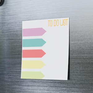 Könnte beinhalten: Ein weißer To-Do-Listen-Notizblock mit farbigen pfeilförmigen Checklistenpunkten ist mit Magneten an einer Edelstahl-Kühlschranktür befestigt. Der Notizblock trägt die orangefarbenen Worte "TO DO LIST".