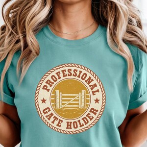 May include: A teal short-sleeve t-shirt featuring a circular graphic with a rope border. The design includes the text "PROFESSIONAL GATE HOLDER" in brown letters, two small stars, and a central illustration of a wooden farm gate on a gold background.