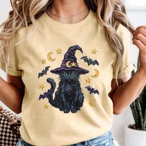 Może przedstawiać: Bladoż&oacute;łty t-shirt z grafiką przedstawiającą czarnego kota w fioletowym kapeluszu czarownicy. Wz&oacute;r zawiera ż&oacute;łte p&oacute;łksiężyce, gwiazdy i małe fioletowe nietoperze otaczające kota. Koszulka ma kr&oacute;tkie rękawy i okrągły dekolt.