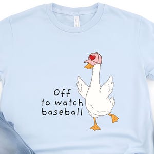 May include: Pale blue t-shirt featuring a cartoon goose wearing a pink baseball cap, with the text "Off to watch baseball". The image also shows blue jeans and white trainers.