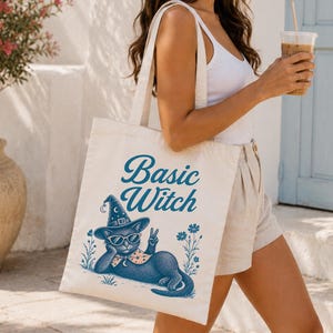 May include: A cream-colored canvas tote bag featuring a blue graphic print of a cat wearing a witch hat and sunglasses, making a peace sign. The text "Basic Witch" is printed in a blue script font above the cat, surrounded by small floral accents.