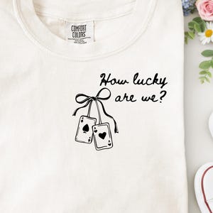 May include: Off-white t-shirt featuring the phrase "How lucky are we?" and a graphic of two playing cards suspended from a bow. The shirt is folded on a white surface, with a pair of jeans and a sweater visible.
