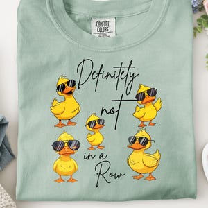 May include: A pale green t-shirt featuring the phrase "Definitely not in a Row" and cartoon ducks in sunglasses. The shirt is displayed on a white surface, with jeans and white trainers visible in the background.
