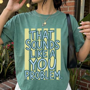 Peut inclure: Un t-shirt à manches courtes vert sauge avec un motif graphique à rayures verticales jaunes et le texte "THAT SOUNDS LIKE A YOU PROBLEM." en lettres bleu clair. Le t-shirt est associé à une jupe plissée blanche et un sac à bandoulière noir.