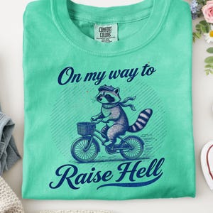 May include: A turquoise t-shirt featuring the phrase "On my way to Raise Hell" and a raccoon on a bicycle. The raccoon wears a hat and scarf. The shirt is next to blue jeans, a cream-coloured sweater, and white trainers.