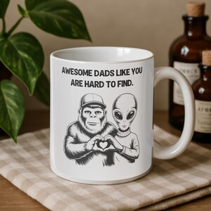May include: White ceramic mug with the text "AWESOME DADS LIKE YOU ARE HARD TO FIND." The mug features a graphic of a Bigfoot and an alien forming a heart shape with their hands. The mug has a large handle and is on a tan and white checkered surface.