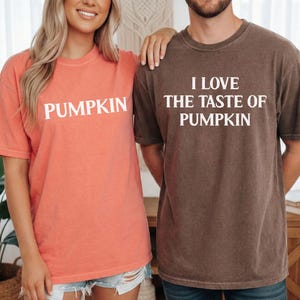 May include: A coral-colored short-sleeve t-shirt with the word PUMPKIN printed in white and a dark brown short-sleeve t-shirt with the white text I LOVE THE TASTE OF PUMPKIN printed on the front. Both shirts feature a crew neckline.