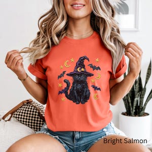 Może przedstawiać: Jaskrawopomarańczowy t-shirt z grafiką czarnego kota w fioletowym kapeluszu czarownicy, otoczonego ż&oacute;łtymi p&oacute;łksiężycami, gwiazdami i czarnymi nietoperzami, umieszczoną na środku klatki piersiowej koszulki z kr&oacute;tkim rękawem i okrągłym dekoltem.