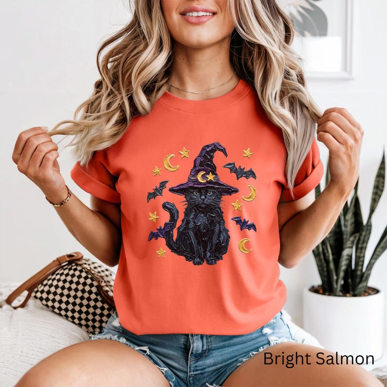 Może przedstawiać: Jaskrawopomarańczowy t-shirt z grafiką czarnego kota w fioletowym kapeluszu czarownicy, otoczonego ż&oacute;łtymi p&oacute;łksiężycami, gwiazdami i czarnymi nietoperzami, umieszczoną na środku klatki piersiowej koszulki z kr&oacute;tkim rękawem i okrągłym dekoltem.