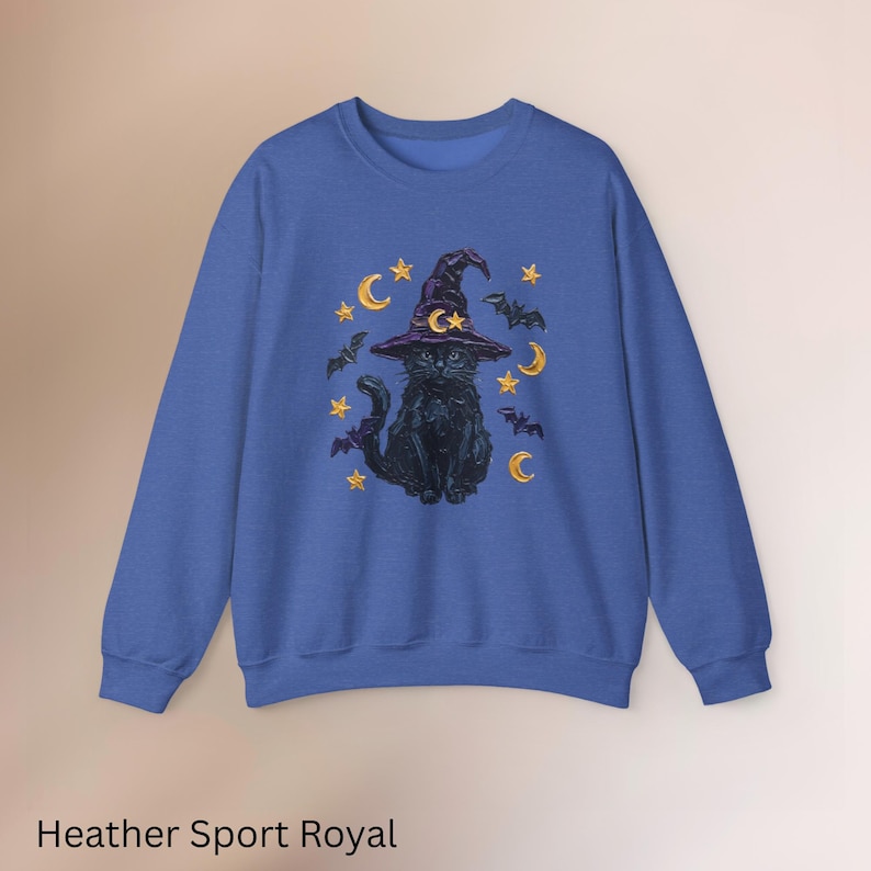 Może przedstawiać: Bluza z okrągłym dekoltem w kolorze heather sport royal z centralną grafiką przedstawiającą czarnego kota w fioletowym kapeluszu czarownicy. Wz&oacute;r otaczają ż&oacute;łte p&oacute;łksiężyce, gwiazdy oraz małe czarne nietoperze tworzące ilustrację.